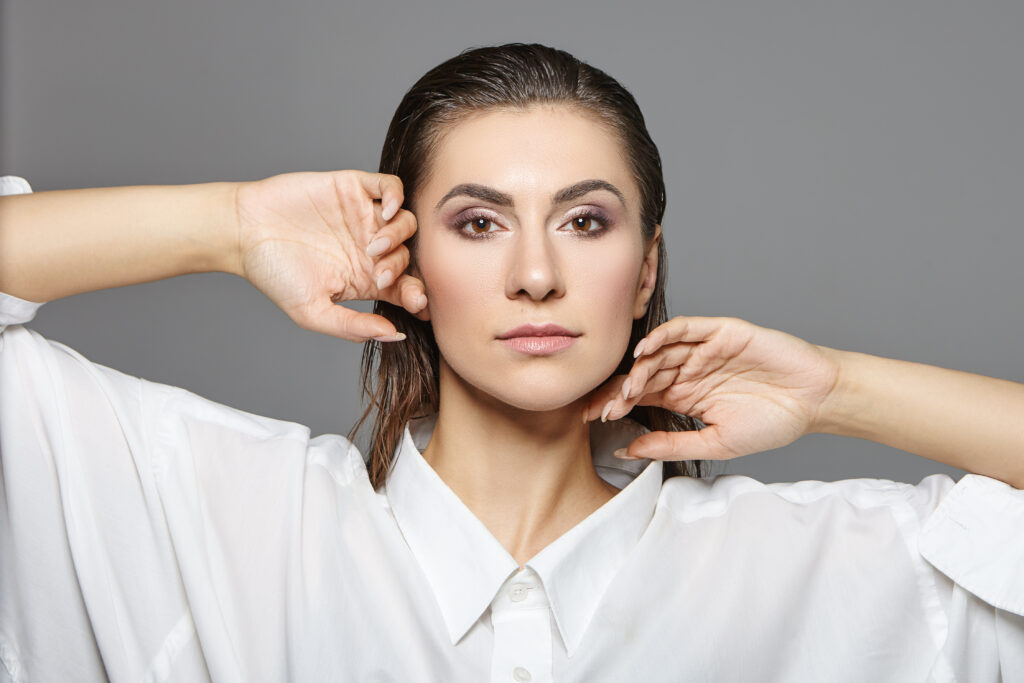 Close-up portrait of a woman with slicked-back hair wearing a white shirt, hands framing her face against a gray backdrop.